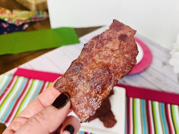 One slice of air fryer bacon being held by an individual wearing black nail polish. White background. 